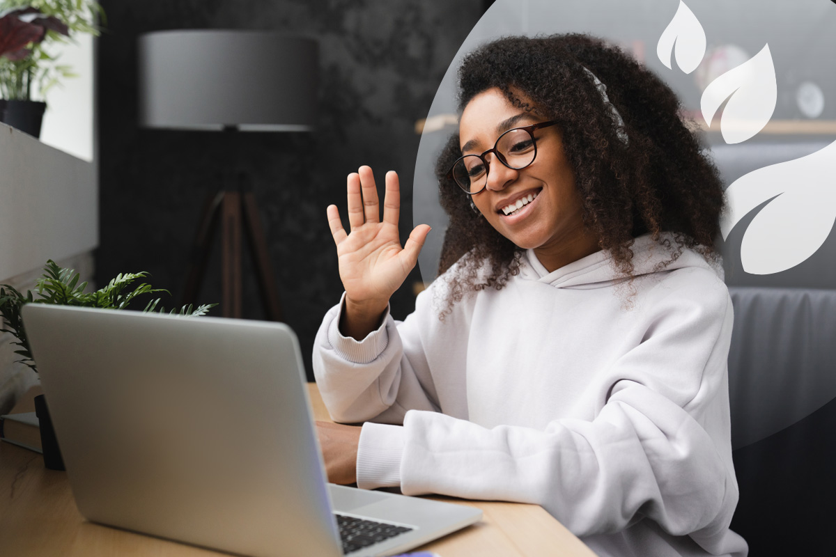 A young person with glasses and headphones, smiling and waving at a laptop screen, engaged in an online video call.