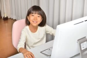 A photograph of a Primary-age girl with black hair sitting at a desktop computer and smiling at the camera.