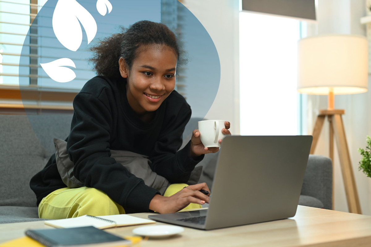 Smiling student with a mug in hand using a laptop at home, seated on a sofa with notebooks on the table.
