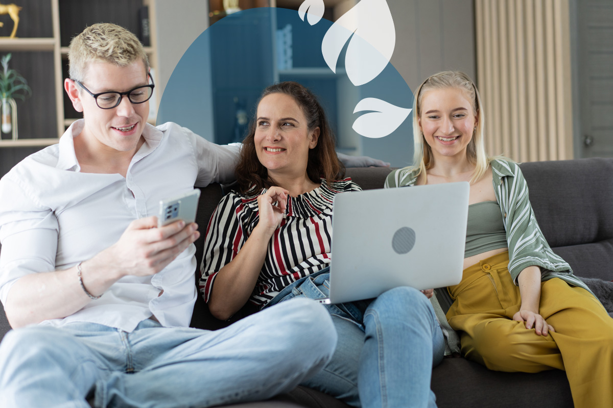 Three people sitting on a sofa smiling while looking at a laptop and phone together