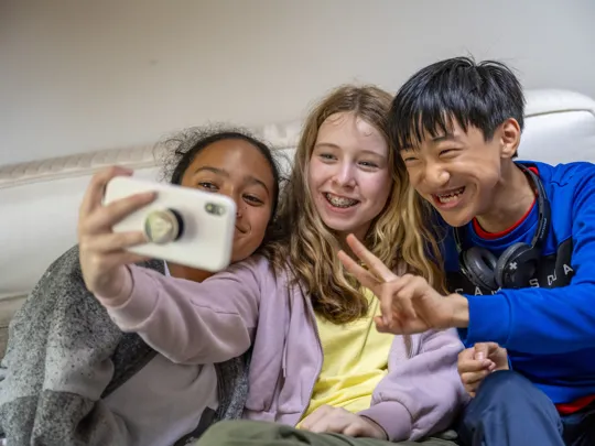 Three smiling teenagers taking a selfie together, sitting close on a sofa.