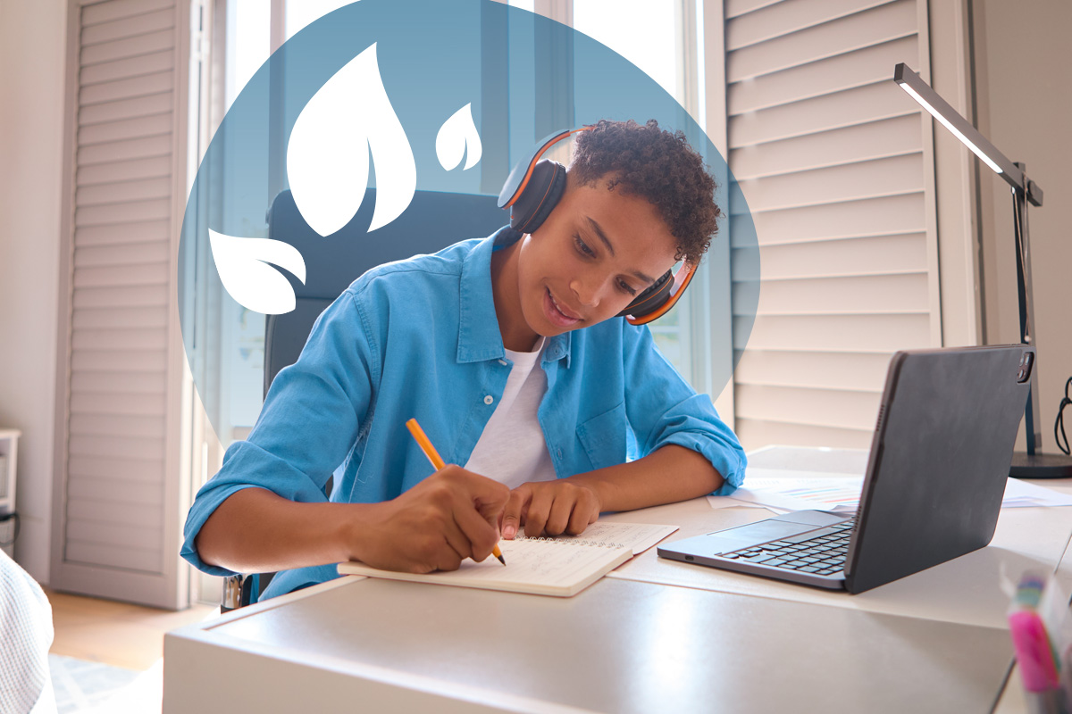 A young person wearing headphones, writing in a notebook at a desk with a laptop and school supplies.