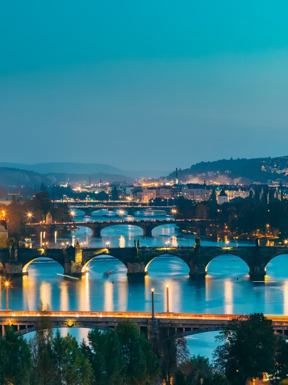 Panoramic twilight view of Prague with iconic bridges and historic architecture lit up.