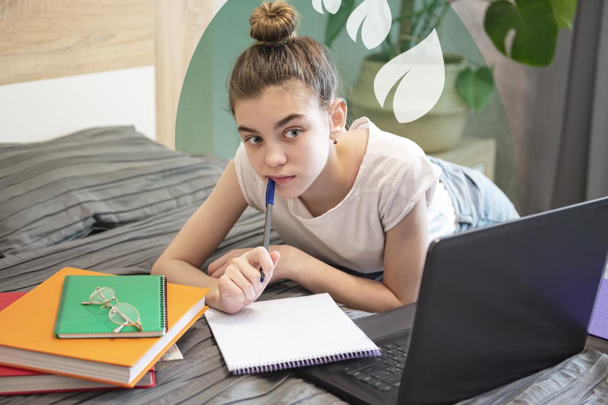 Teen girl lying on bed, thoughtfully holding a pen while studying with laptop and folders.