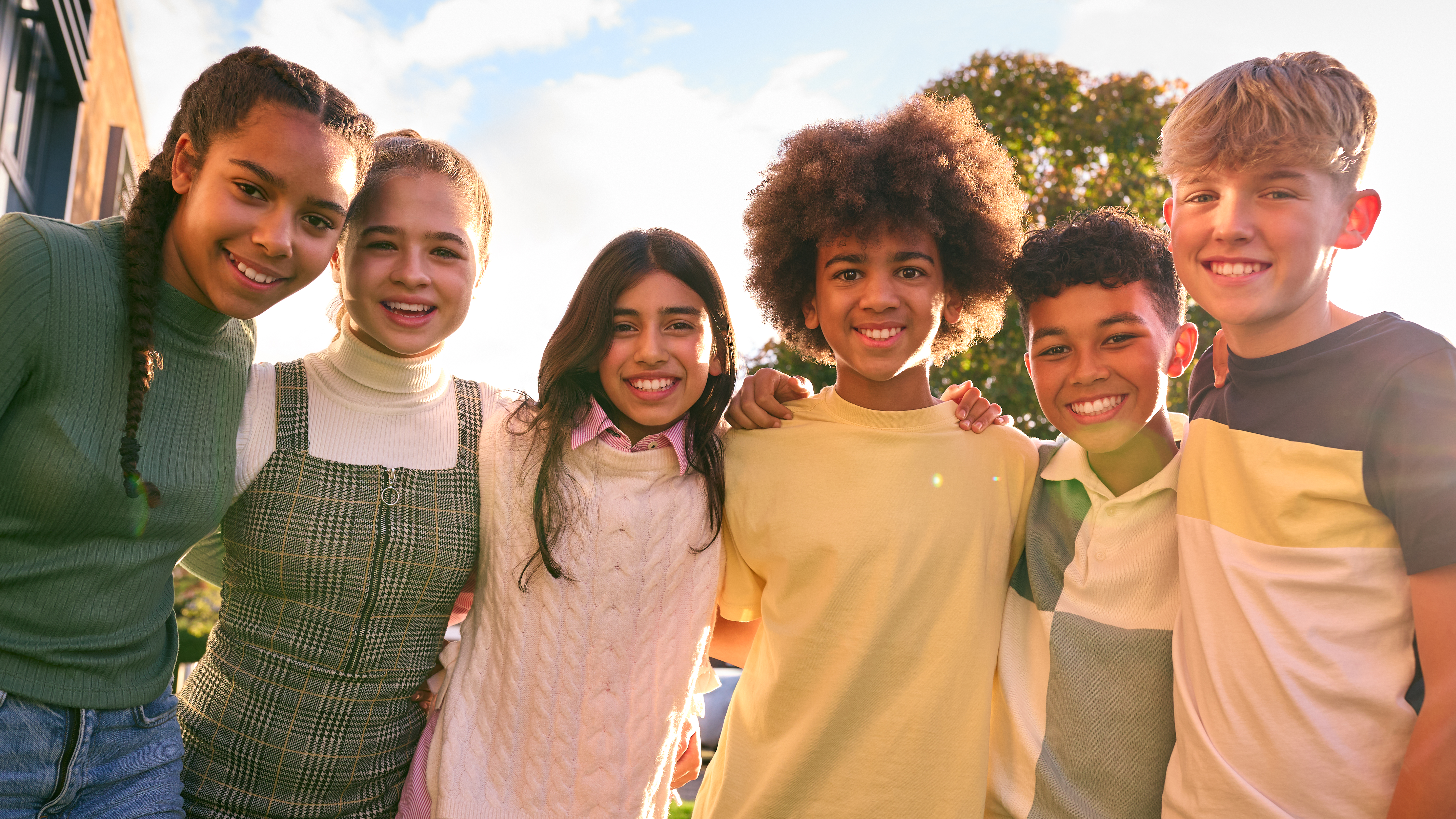 Group of six smiling children standing close together outdoors on a sunny day.