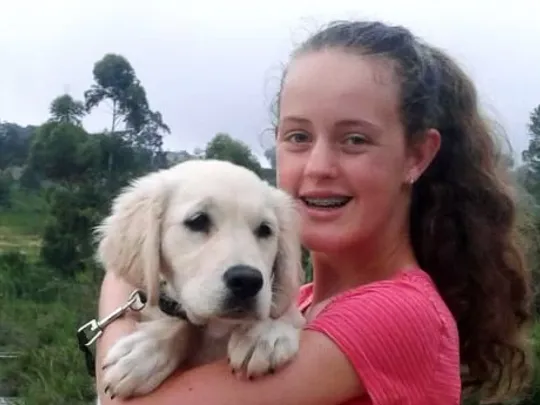 A young woman smiling and holding a light-coloured puppy, standing in a natural landscape with trees and water in the background.