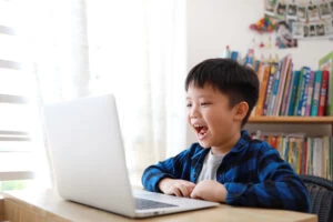 A photograph of a very young boy with short black hair smiling at a laptop computer whilst sitting at a table.