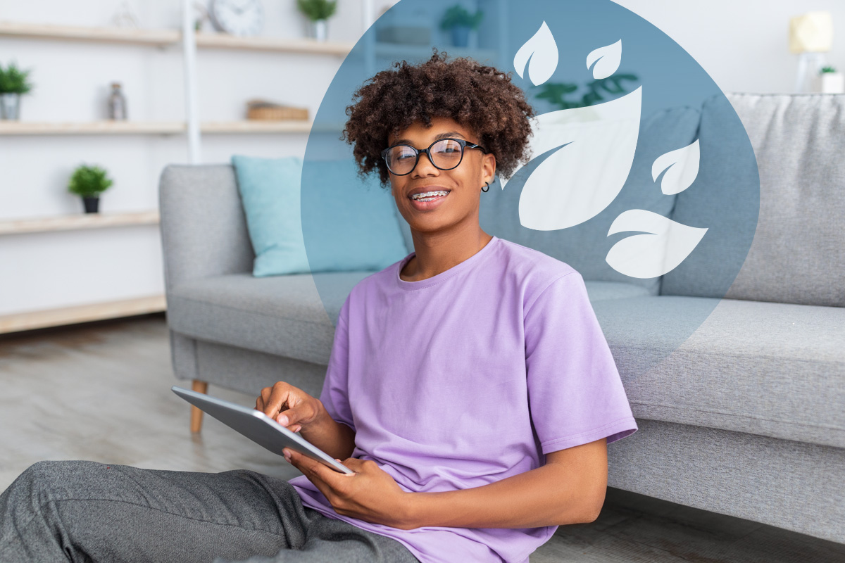 A teenager with glasses and braces, smiling and looking at the camera while holding a tablet, sitting on the floor in a casual setting.