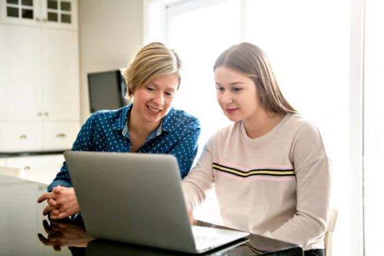 A homeschooling mother and a daughter looking at a laptop at their kitchen table.
