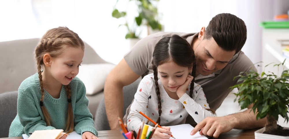A dad homeschooling his two young daughters at a desk.
