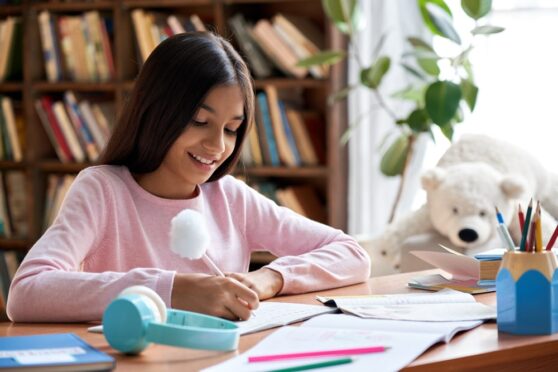 A homeschooling young female student writing in an exercise book at her bedroom desk.