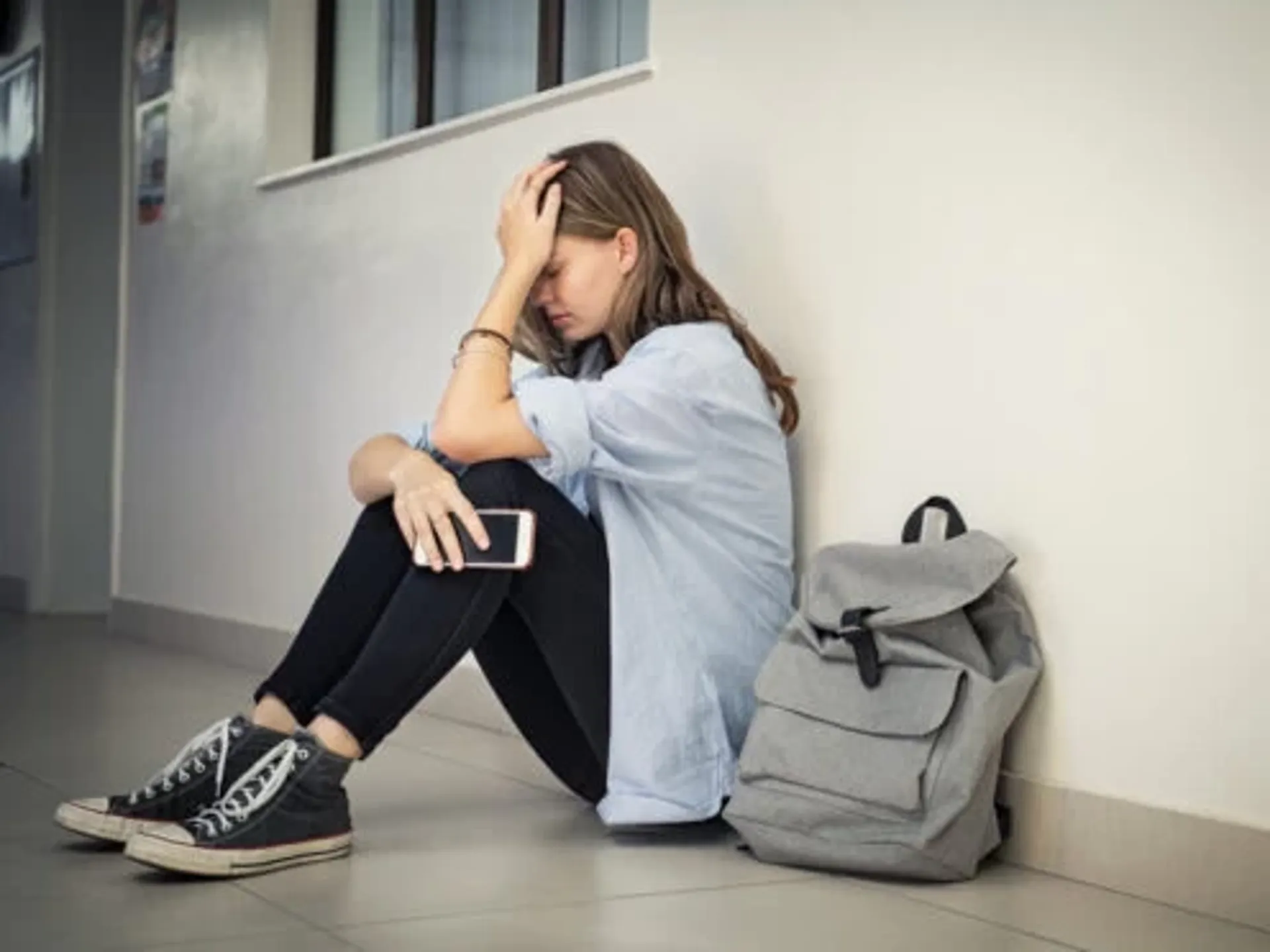 A young girl sitting against a wall with her head in her hand, and her phone in the other. She is looking anxious and upset.