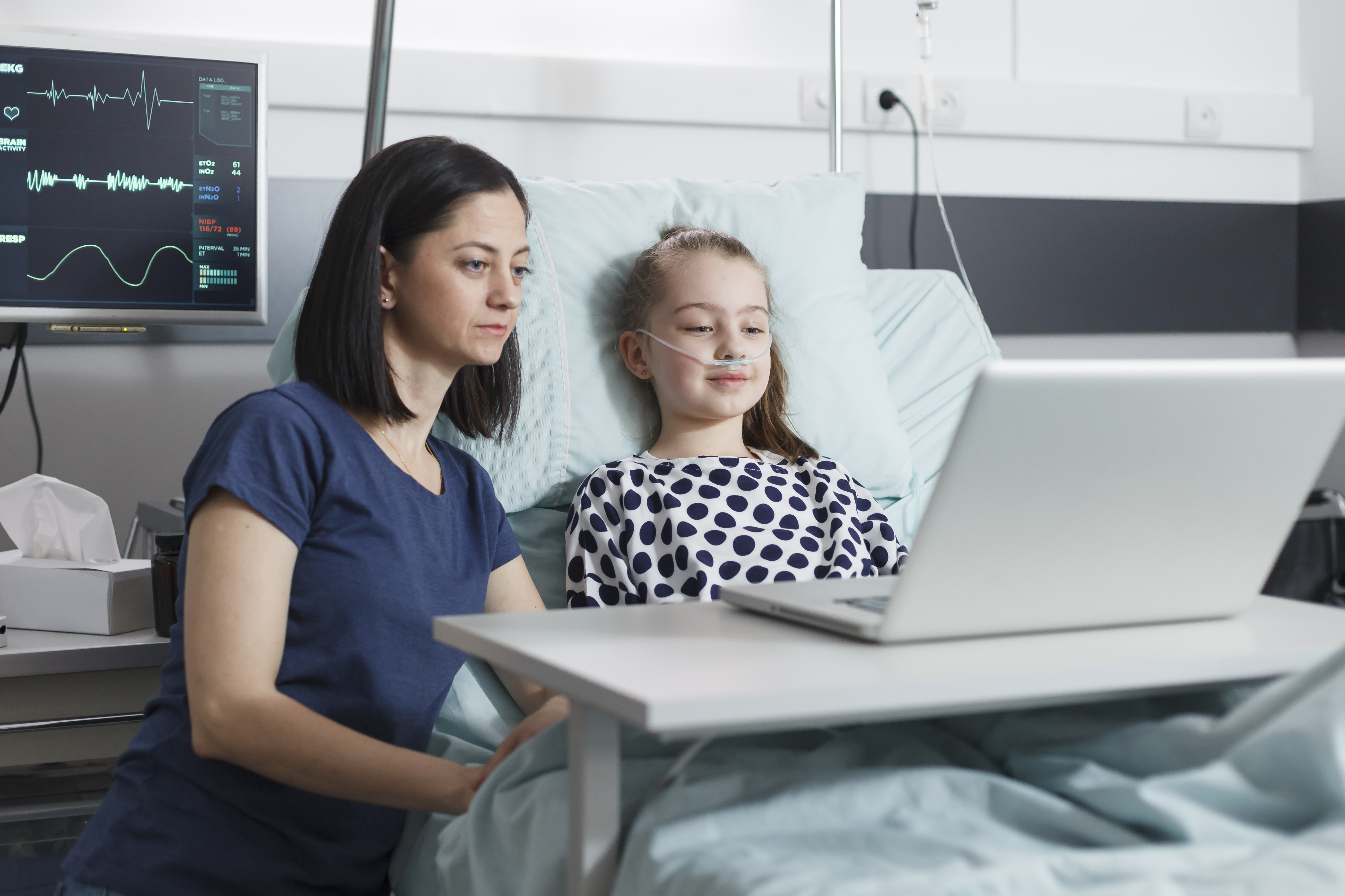 Girl recovering in hospital using a laptop with supportive adult by her side