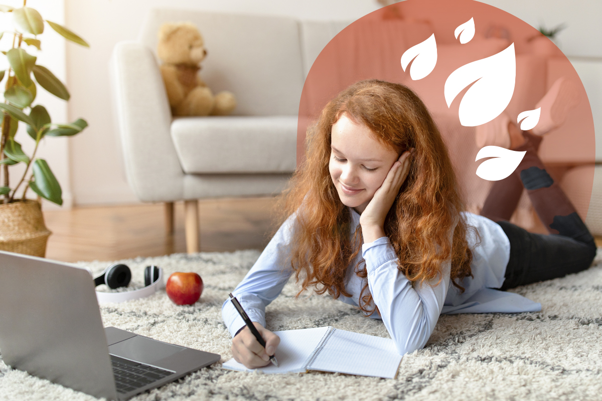 Smiling girl lying on rug, writing in a notebook with a laptop and apple nearby.