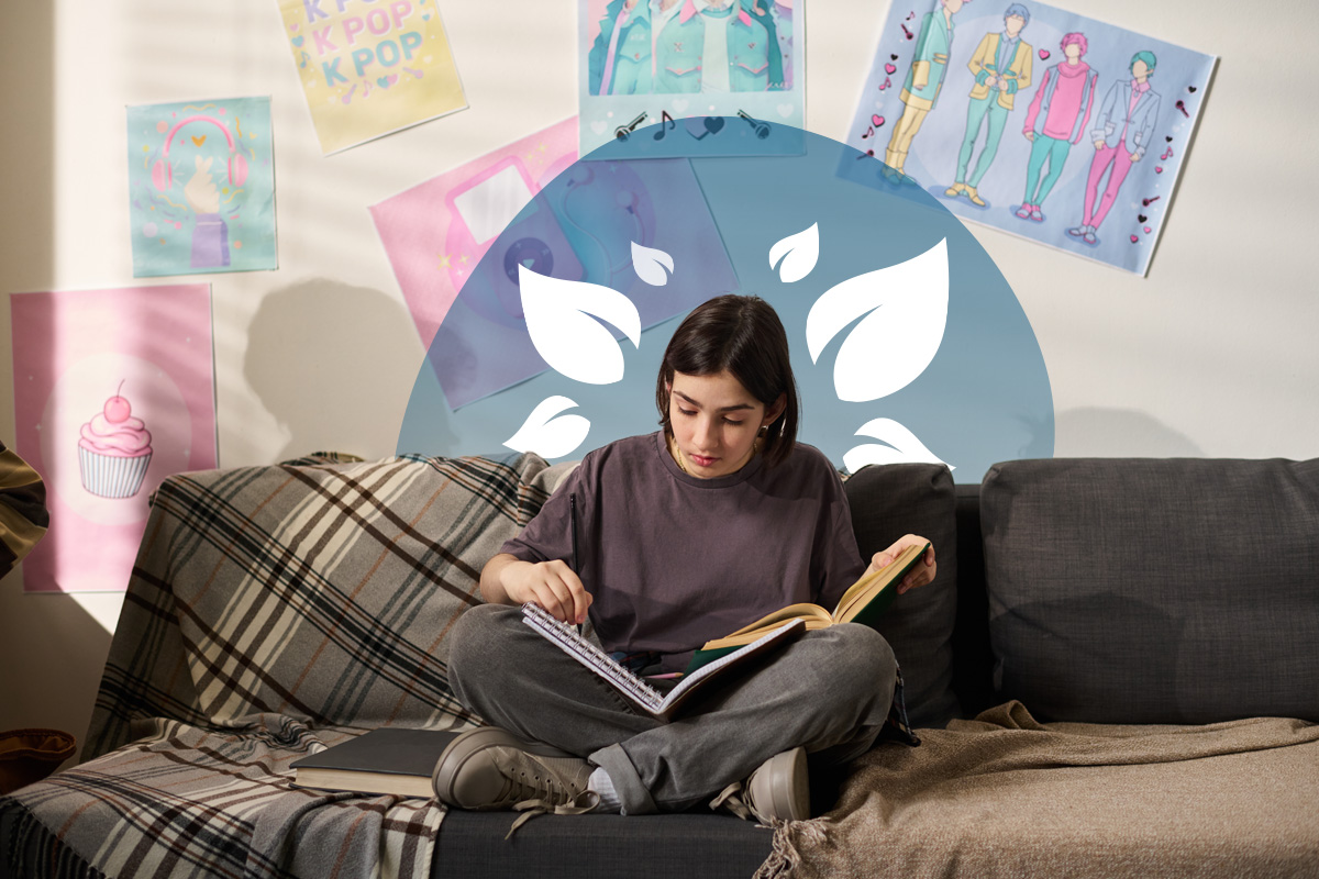  A young person with dark hair sits cross-legged on a patterned couch, engrossed in reading an open book. Various posters, including 
