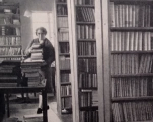 An old black and white photograph of a female library worker in a library. The lady is carrying a big pile of books and walking towards the camera.