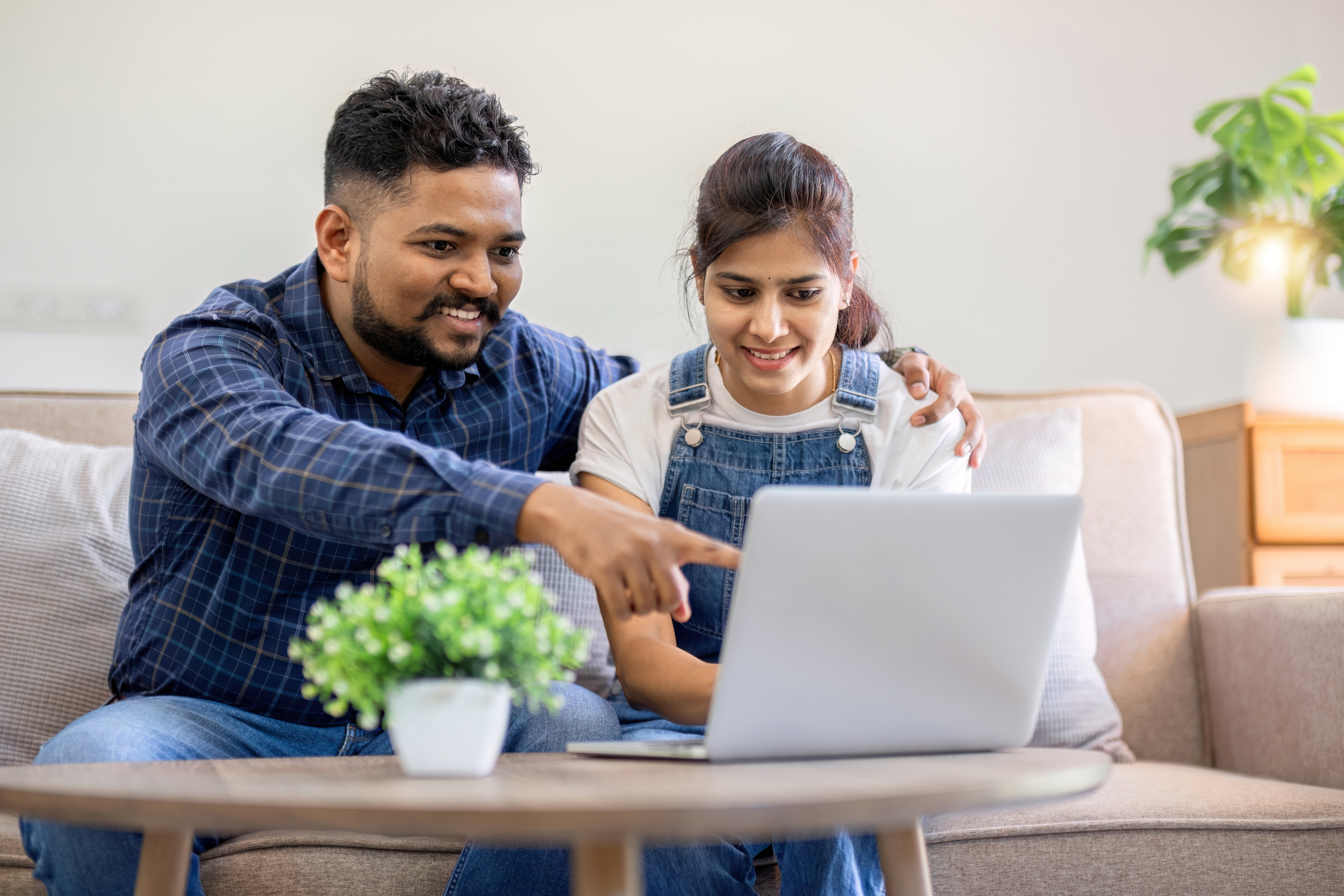Smiling couple sitting on a sofa looking at a laptop together, with the man pointing at the screen.