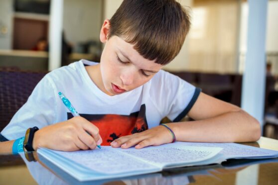A homeschooling young boy with brown hair writing in a notepad at his desk.