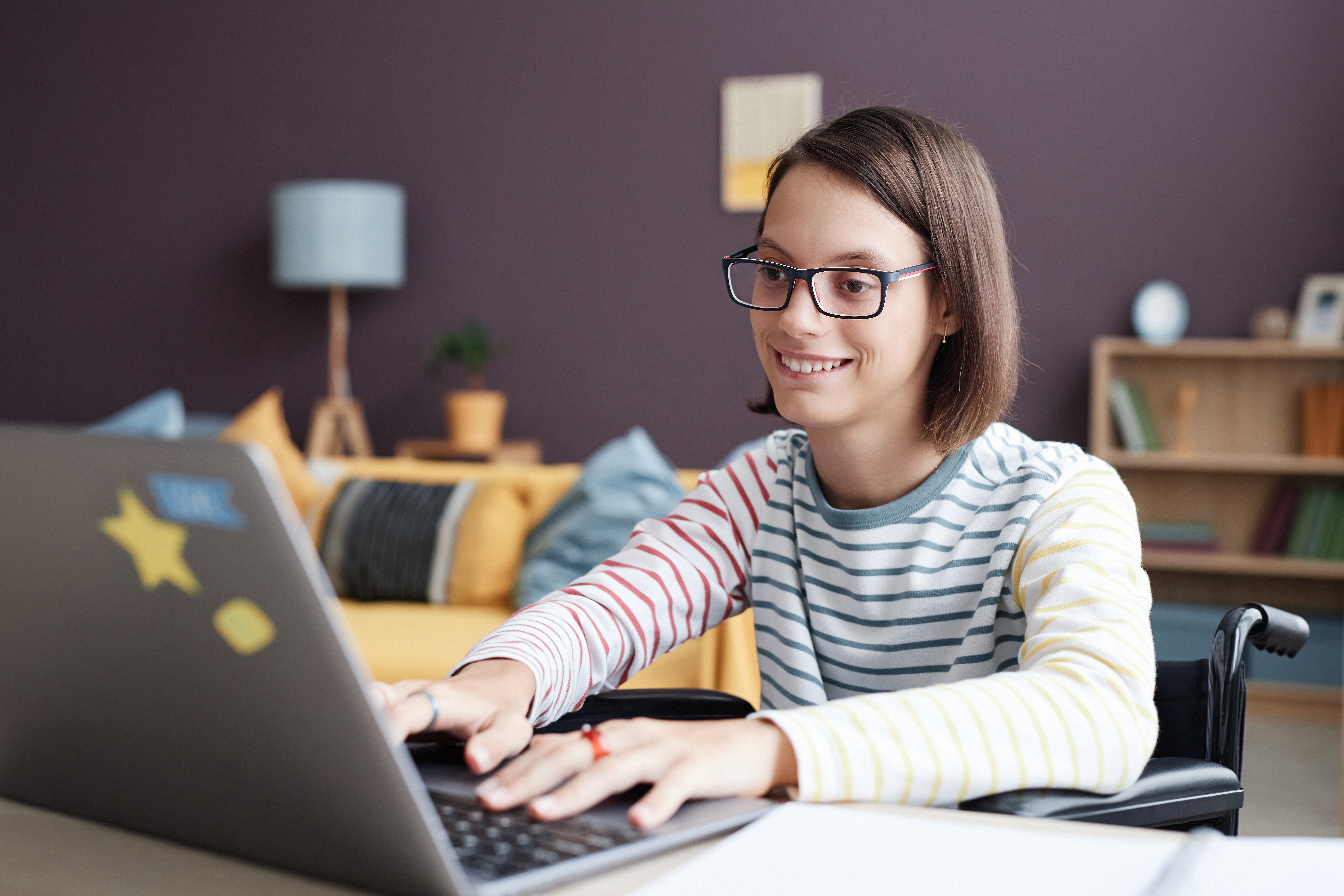 Teenage girl studying on laptop in wheelchair in comfortable home setting
