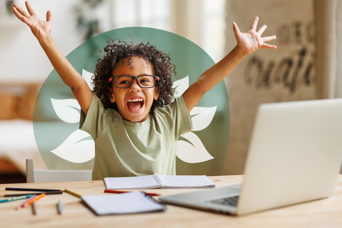 A young student with glasses, smiling excitedly with arms raised, sitting at a desk with a laptop and open notebook.