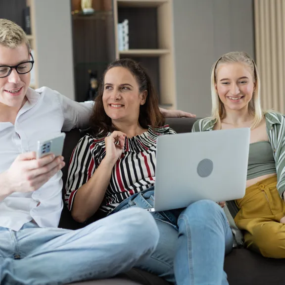 Three people sitting on a sofa smiling while looking at a laptop and phone together