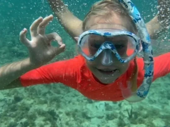 A child wearing a snorkel mask and orange top underwater, giving an