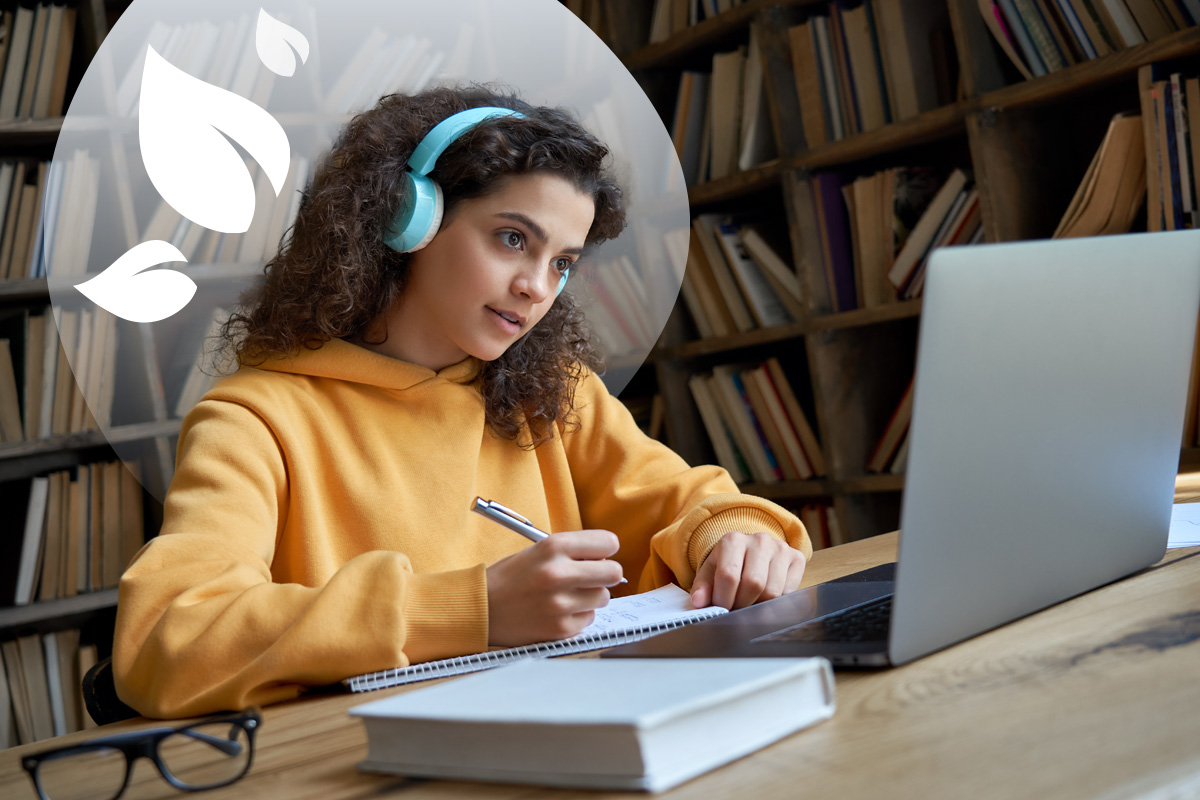 A student with curly hair wearing headphones, writing in a notebook while looking at a laptop, with a blurred bookshelf background and a green leafy design.