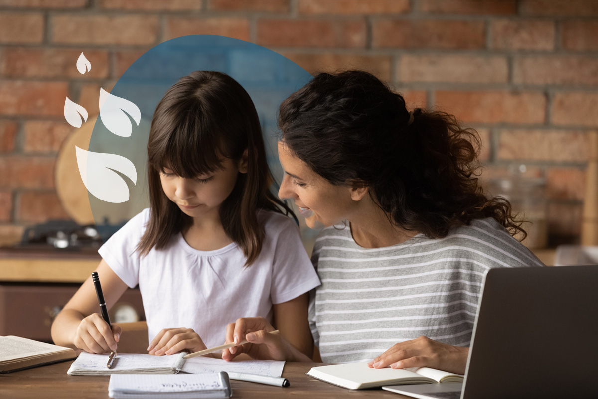 An adult smiling and assisting a young person who is writing in a notebook at a wooden table, with a laptop nearby.