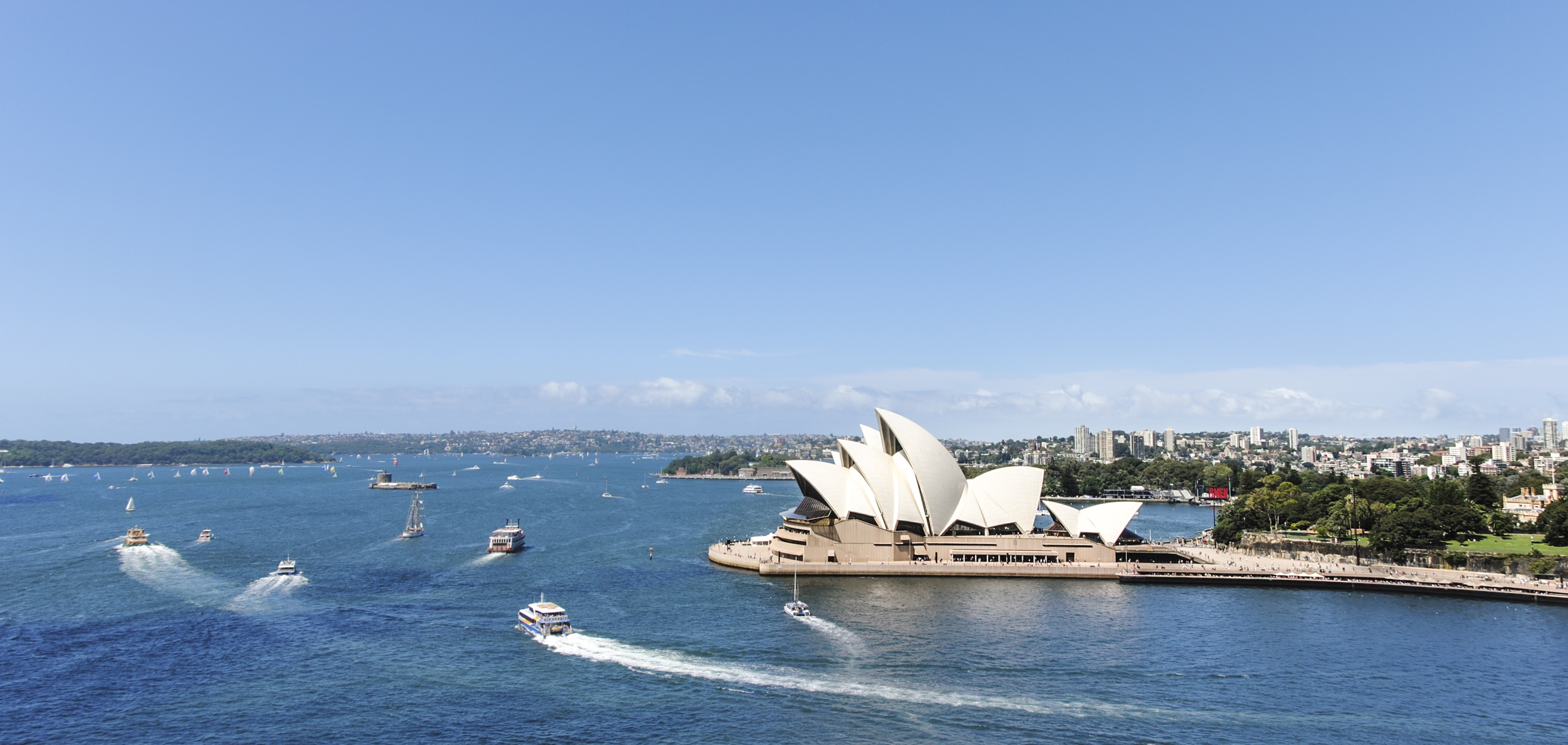 Sydney Opera House beside the harbour with boats leaving white trails in the deep blue water