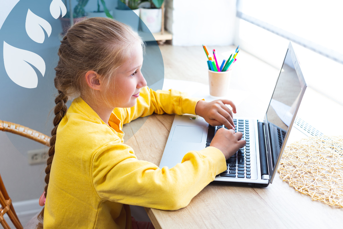 A young student with braided hair, wearing a yellow top, looking at a laptop and typing at a wooden desk.