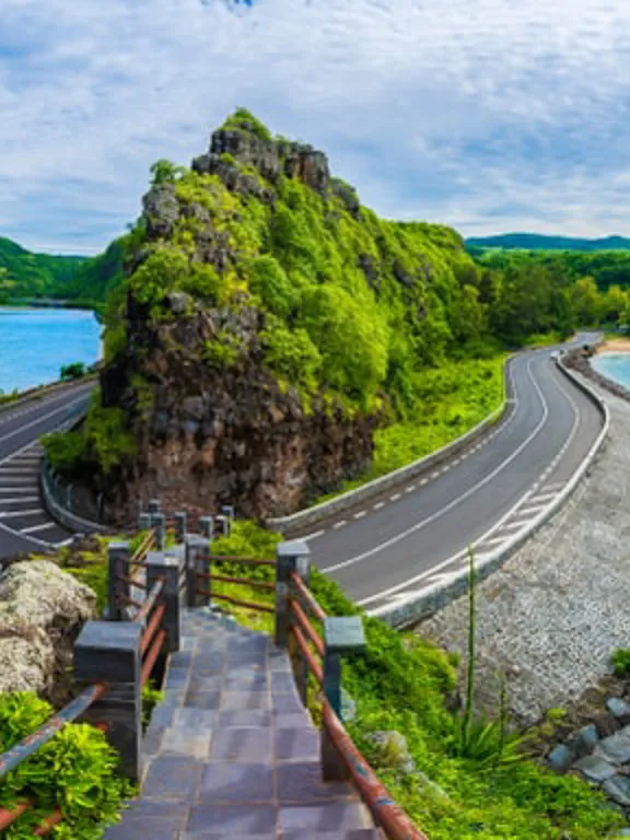 A landscape view of a Mauritian road curving around an outcrop of land over crystal blue sea waters.
