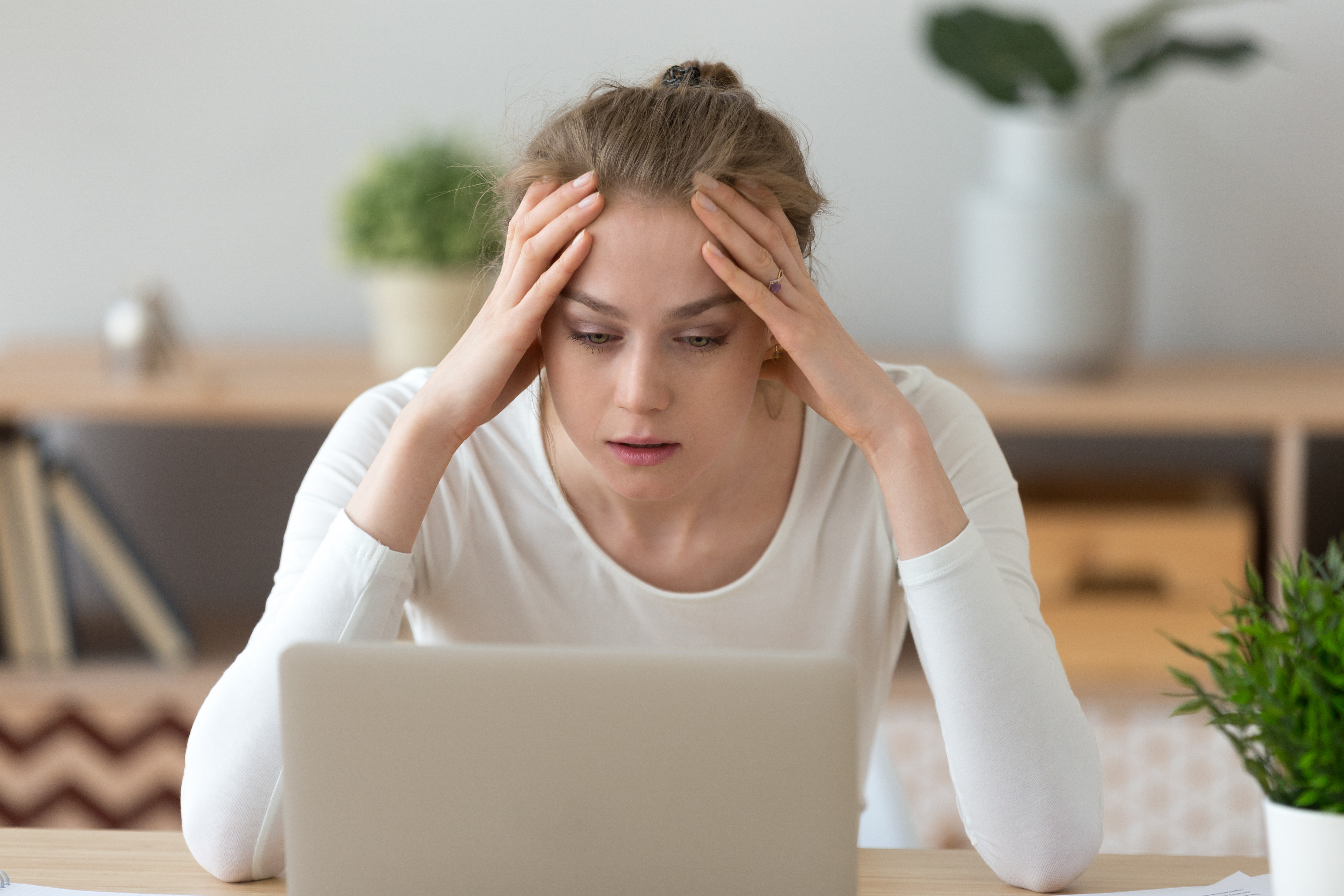 A stressed and anxious secondary student holds her head in her hands while looking at her laptop screen, representing anxiety.