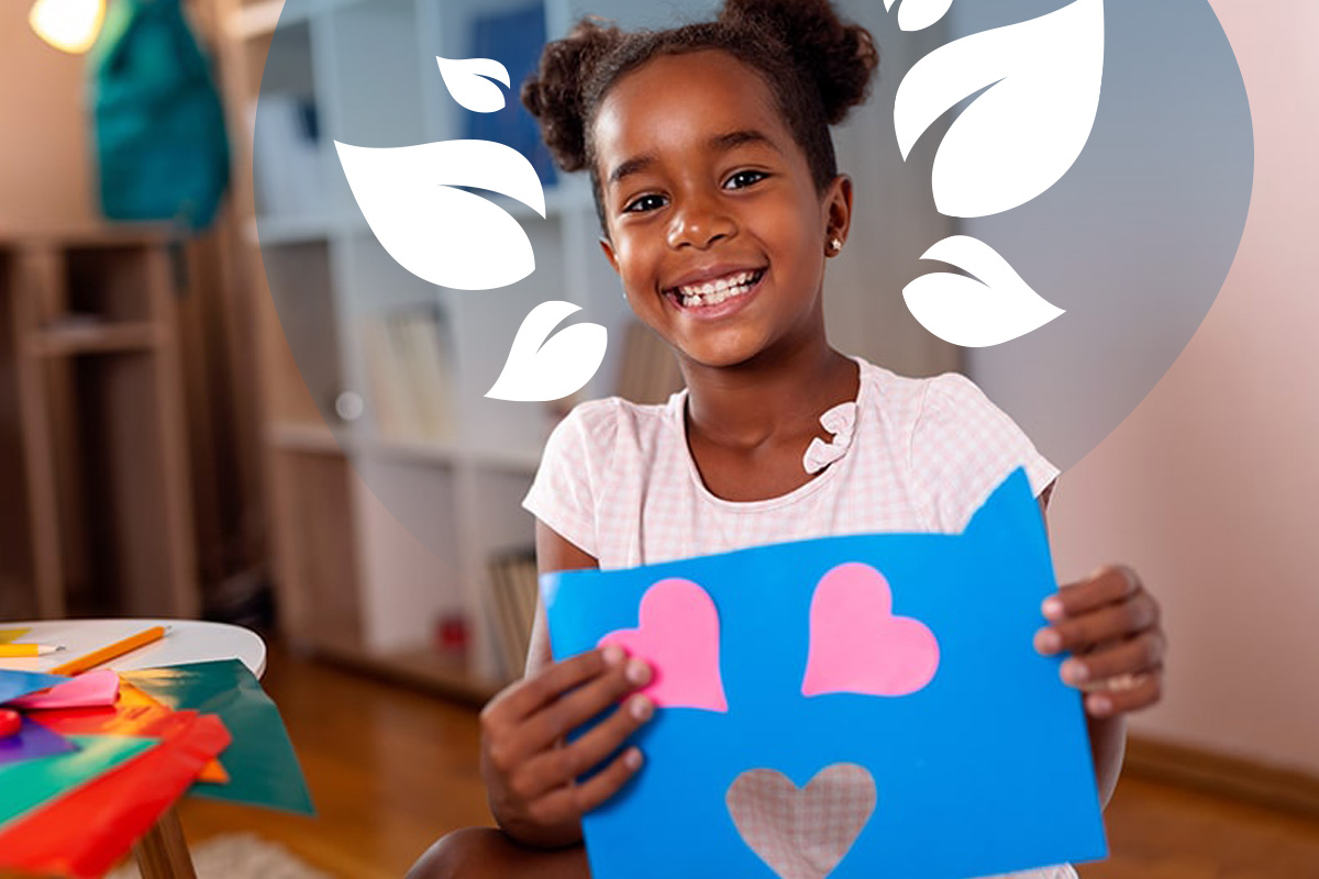 A homeschooling young girl smiling holding up a creative craft picture with pink hearts on blue card.