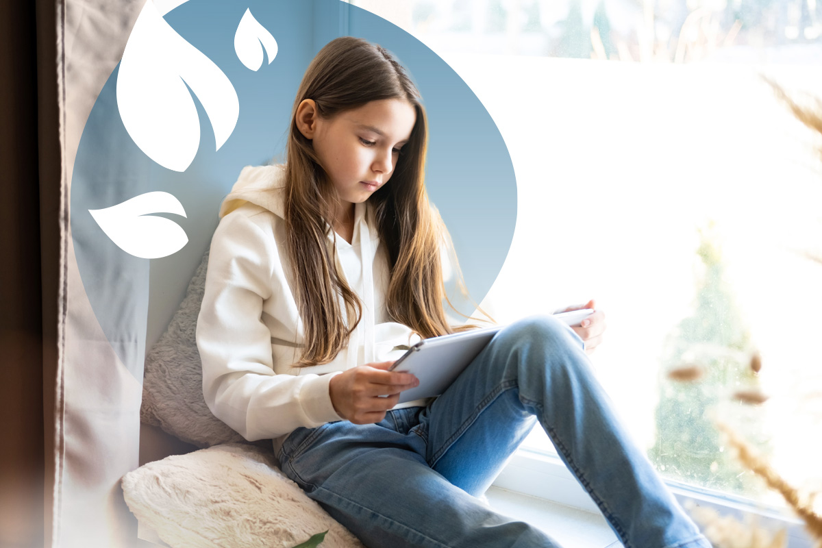 Young girl with long hair reading on a tablet while sitting on a cushion by a bright window.