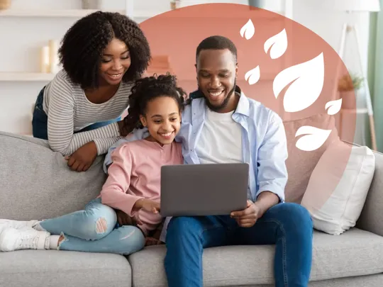 A homeschooling mum, dad and young daughter in their living room smiling at a laptop. The dad and daughter are sat on the sofa, whilst the mother is leaning on the back of it.