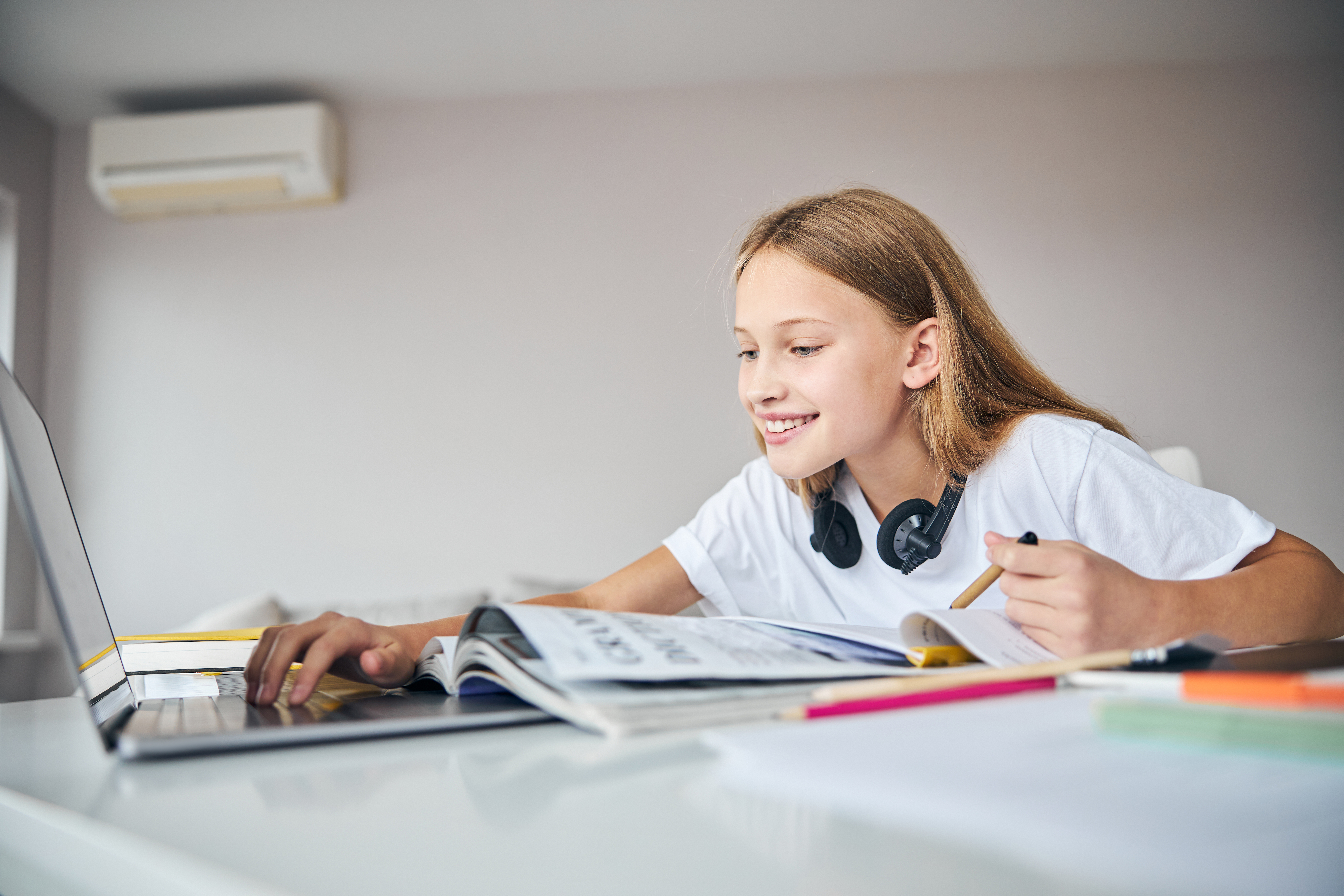 A young girl with headphones smiles while using a laptop to study at her desk, with books and pencils in front of her, representing online education.