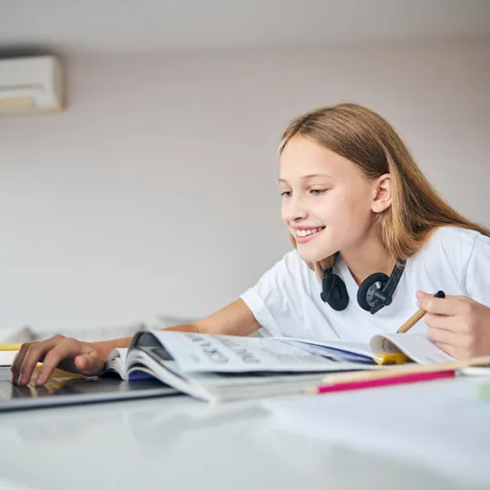 A young girl with headphones smiles while using a laptop to study at her desk, with books and pencils in front of her, representing online education.
