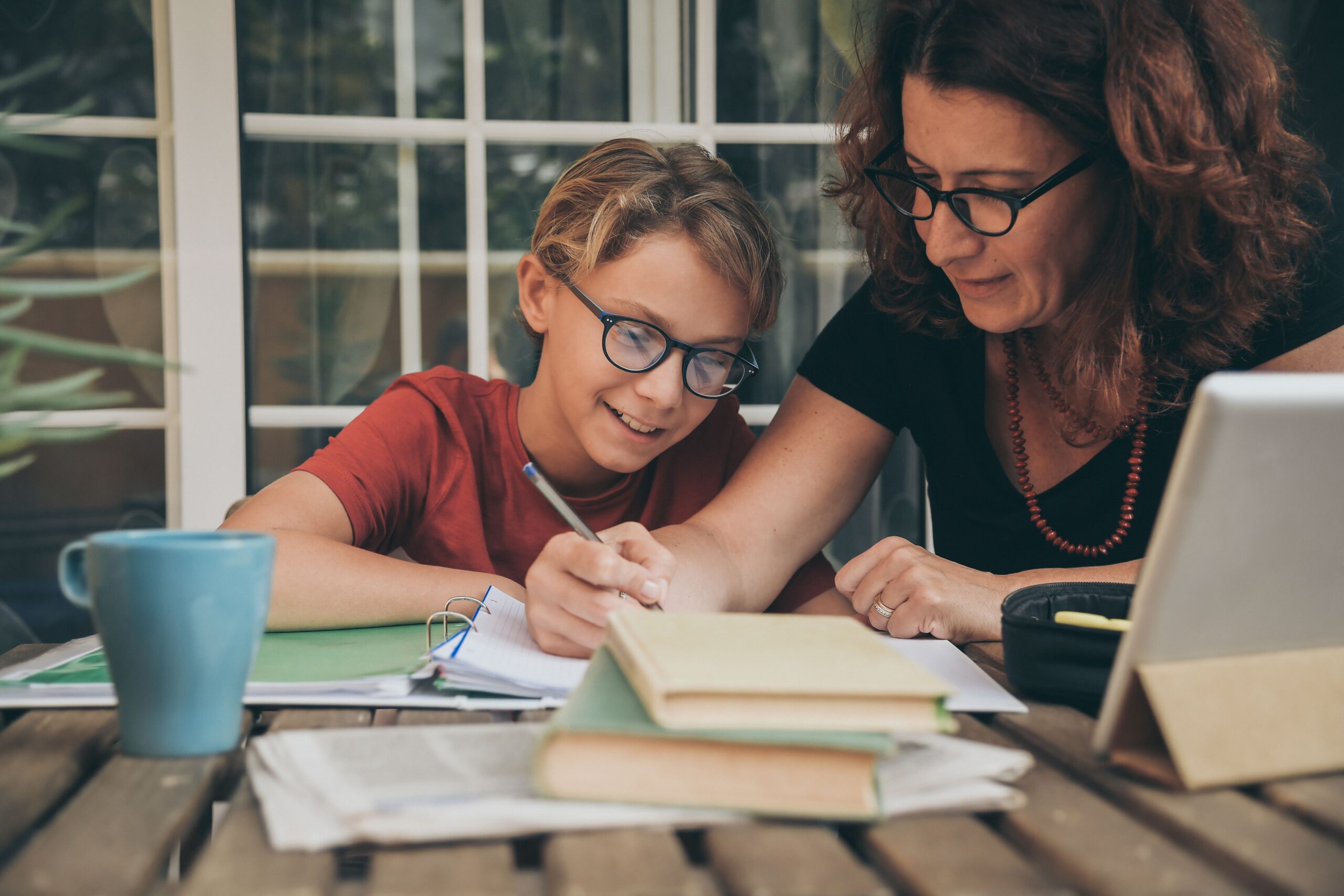 A parent assists a smiling child with their homeschool studies at a table with books and a laptop, illustrating personalized education options for families homeschooling in Riyadh.