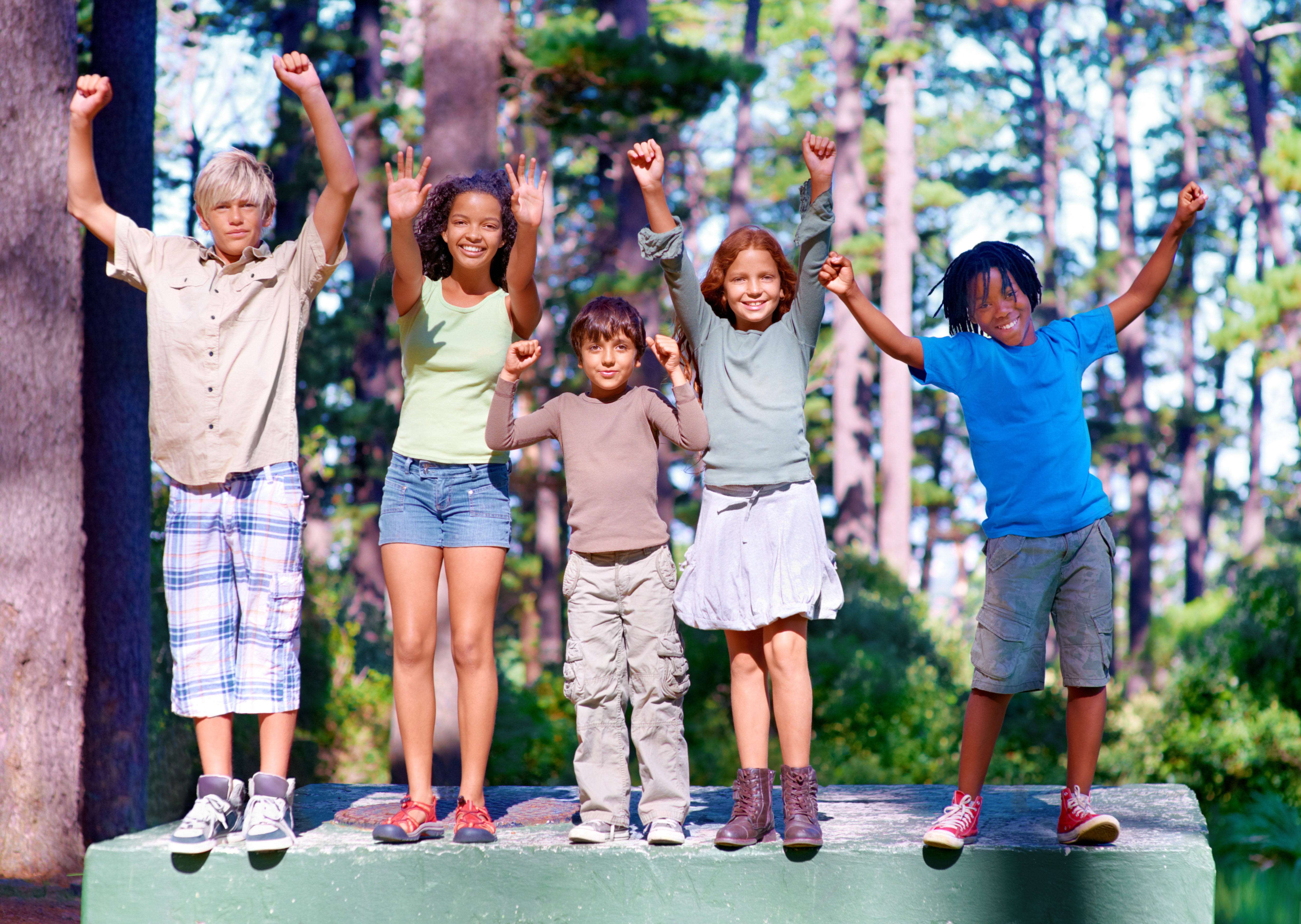 Five children standing with arms raised, smiling in a wooded outdoor setting.
