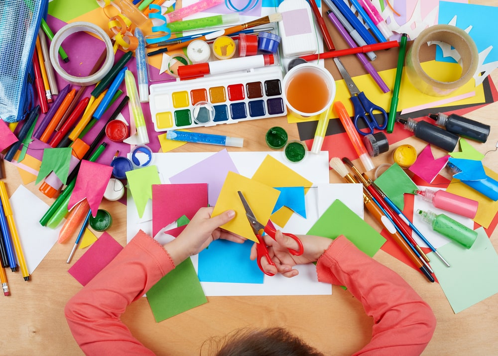 Overhead view of a child using scissors for a colourful paper craft activity surrounded by art supplies