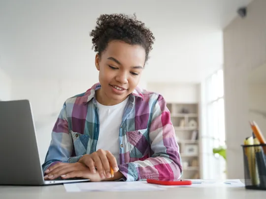 A young student in a plaid shirt sits at a desk, focused on their laptop screen while holding a pencil, engaged in online learning in a bright room.