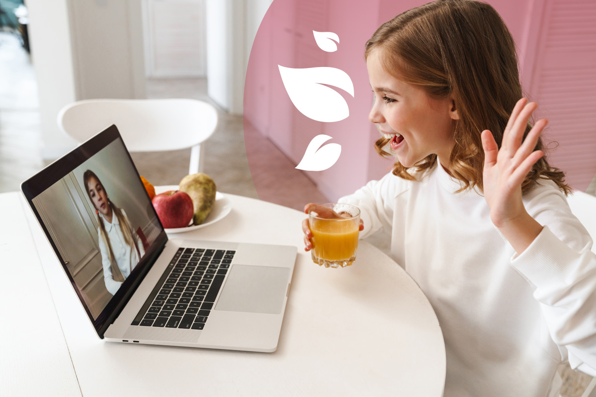 Smiling girl waving at a laptop screen during a video call, with a glass of juice in hand.