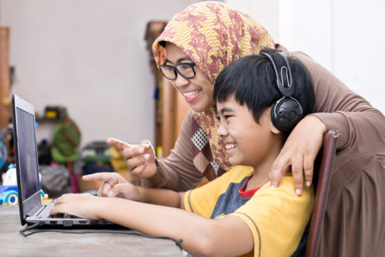A mother helping her young son homeschool. The son is sitting at his desk with his laptop and headphones on, and they are both smiling and pointing at the screen.