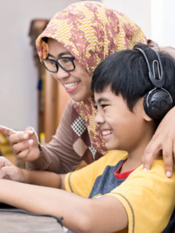 A mother helping her young son homeschool. The son is sitting at his desk with his laptop and headphones on, and they are both smiling and pointing at the screen.