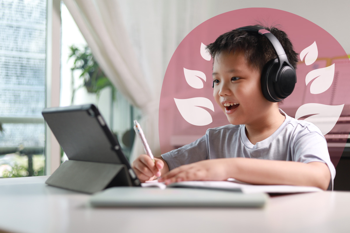 A young student wearing headphones, smiling and writing in a notebook while looking at a tablet, indicating active online learning.