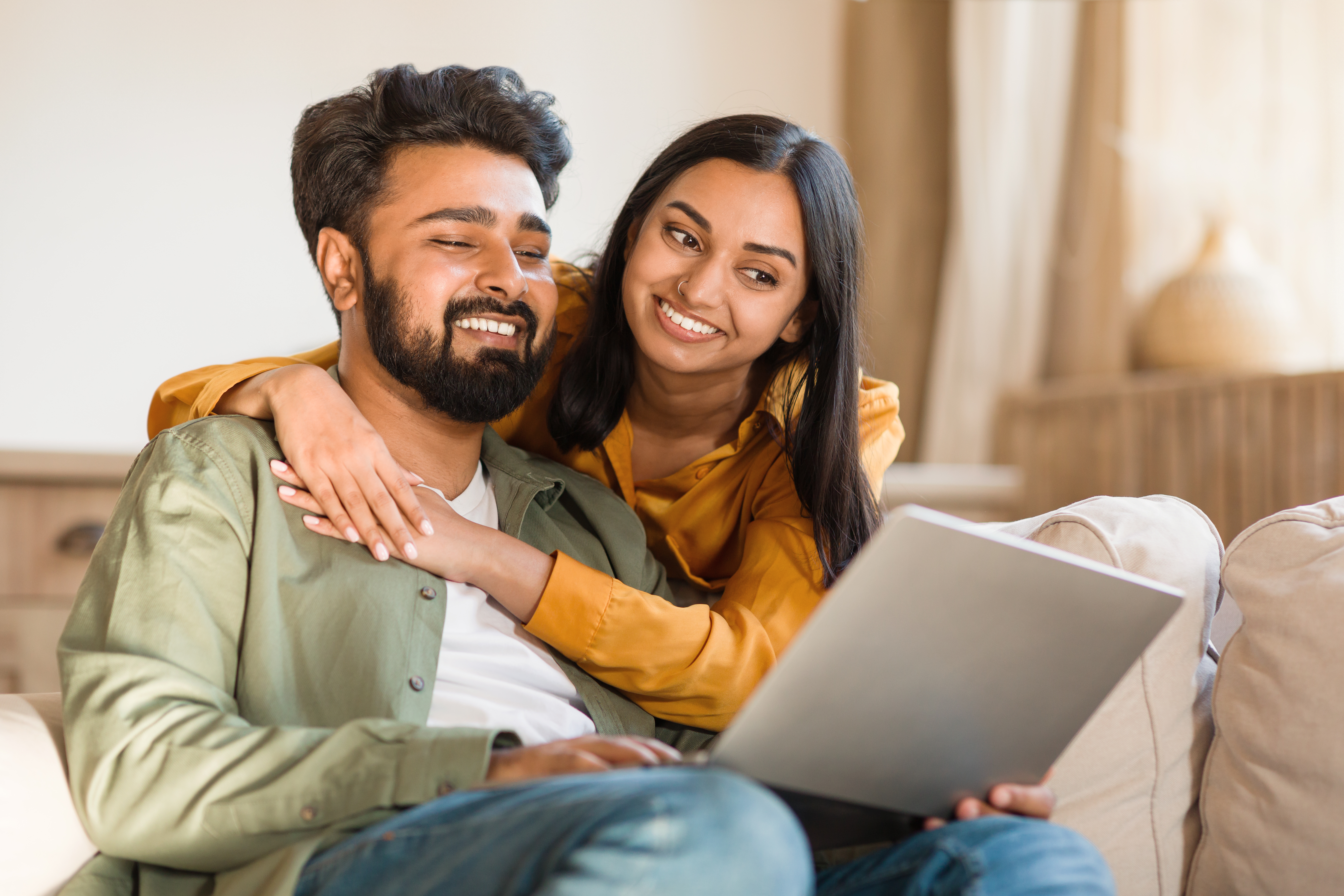 Couple smiling warmly while looking at a laptop together on a cosy living room sofa.