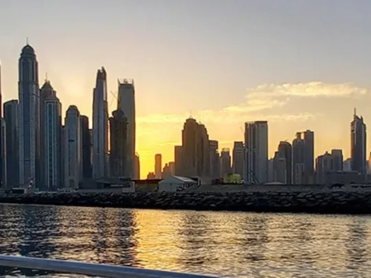 A panoramic view of a city skyline at sunset, with tall buildings reflecting in the water of a harbour or bay.