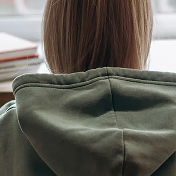 The back of a student wearing a grey hoodie, sitting at a desk with books, a globe, and stationery, looking towards a window.