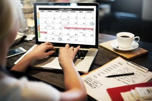 A photograph of a female homeschooling student with her back to the camera working on a laptop, which is displaying a calendar schedule. The student's paper assignment and pen are on the desk next to the laptop.