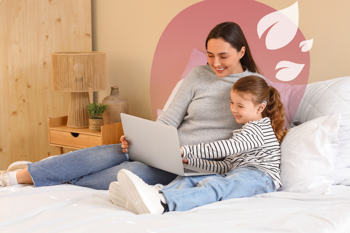  A woman and a child are comfortably seated on a bed, both smiling and looking at a laptop together. The woman is wearing a grey sweater and jeans, while the child is in a striped long-sleeve shirt and jeans. A nightstand with a lamp is visible to the left. A translucent pink overlay with white leaf designs is in the top right.